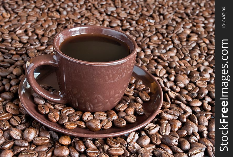 A cup of coffee and matching saucer is surrounded by whole coffee beans. A cup of coffee and matching saucer is surrounded by whole coffee beans