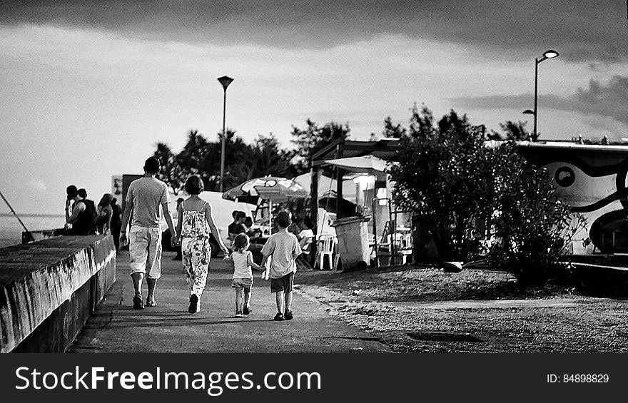 Family, mother, father and two children holding hands and walking at the seaside with boys fishing off the sea wall and a cafe and fairground style entertainment on the right. Family, mother, father and two children holding hands and walking at the seaside with boys fishing off the sea wall and a cafe and fairground style entertainment on the right.