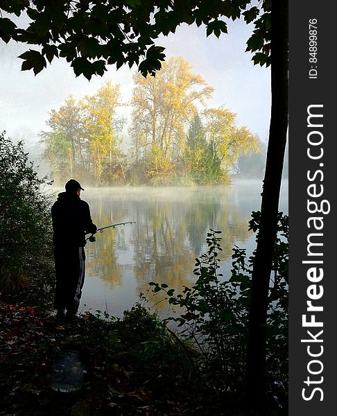 Silhouette of fisherman fishing in tranquil lake or river framed by tree and bushes. A magical sunlit island in the river gives 3D impact. Silhouette of fisherman fishing in tranquil lake or river framed by tree and bushes. A magical sunlit island in the river gives 3D impact.