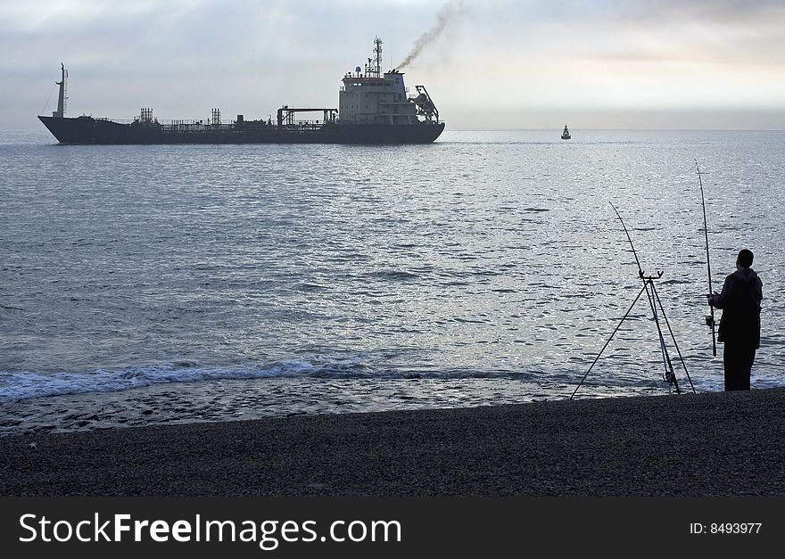 A ship is passing during the sunset while two men are fishing in the sea.