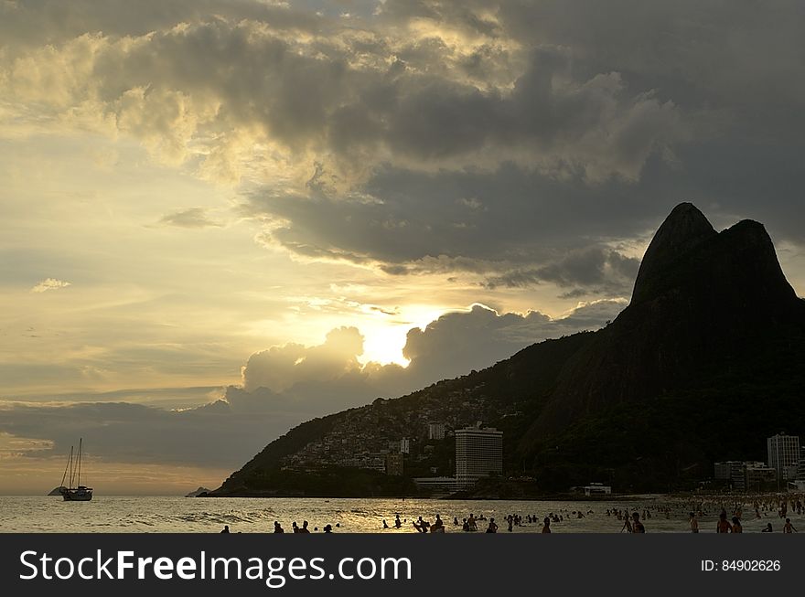 Sailboats off rocky cliff along coastline at sunset in cloudy skies.