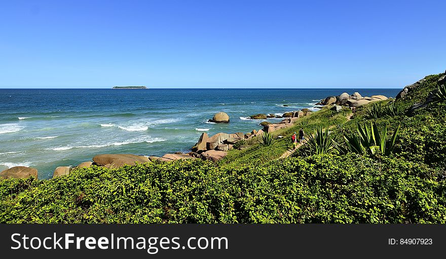 Green hillside down to rocks along blue waters of shore against blue skies on sunny day. Green hillside down to rocks along blue waters of shore against blue skies on sunny day.