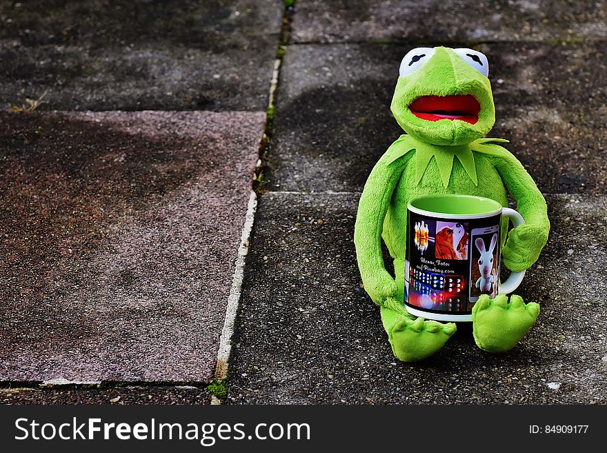 A cute toy frog sitting on the concrete holding a tea mug. A cute toy frog sitting on the concrete holding a tea mug.