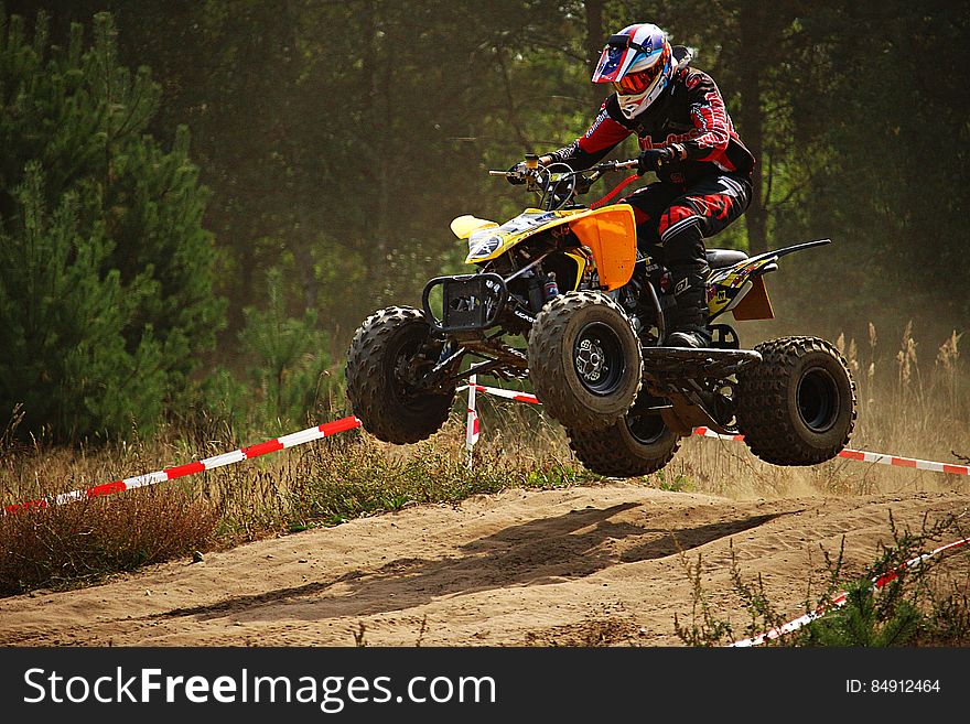 Quad bike with rider in full helmet and black and red suit taking off at hump in the race track throwing up dust, background of green forest. Quad bike with rider in full helmet and black and red suit taking off at hump in the race track throwing up dust, background of green forest.