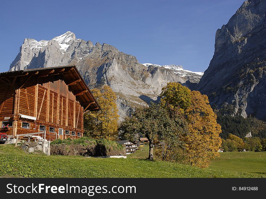 Wooden chalet with exposed beams, and manicured garden with trees and towering over the roof a range of high mountains and blue sky. Wooden chalet with exposed beams, and manicured garden with trees and towering over the roof a range of high mountains and blue sky.