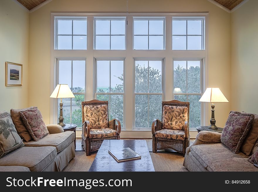 Sitting room in modern apartment with large windows looking onto the garden, vaulted ceiling and with two settees and two arm chairs, coffee table and reading lamps.