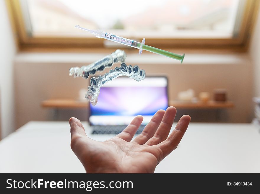 A close up of a hand catching falling denture and syringe and a laptop in the background.