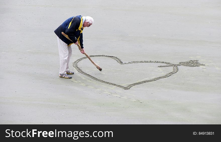 Man Wearing Blue Jacket Holding a Brown Stick Towards the Heart Drawn on Sand