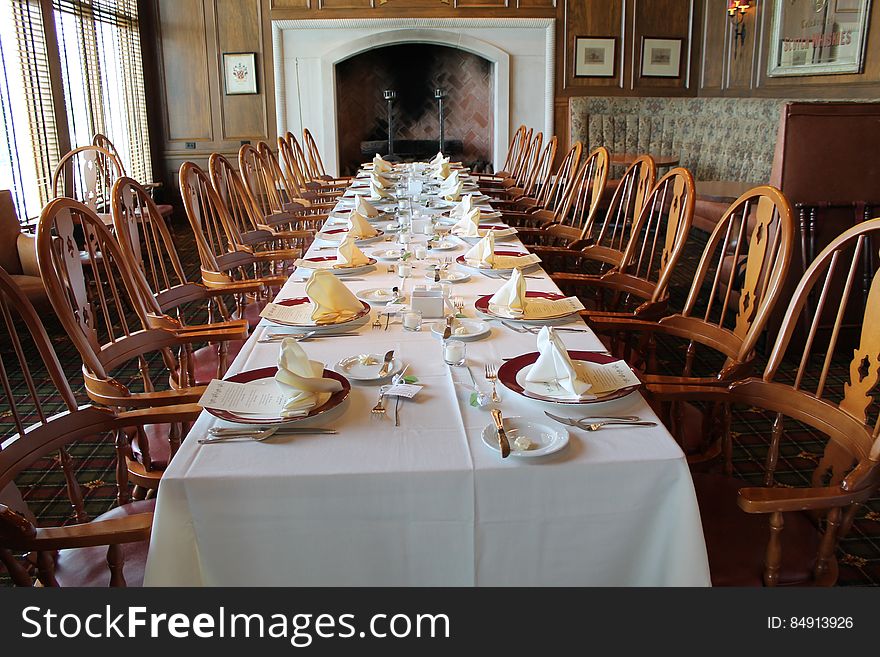 A long dining room table with a white tablecloth and formal place settings, ten chairs on each side, ending in front of a large fireplace. A long dining room table with a white tablecloth and formal place settings, ten chairs on each side, ending in front of a large fireplace.