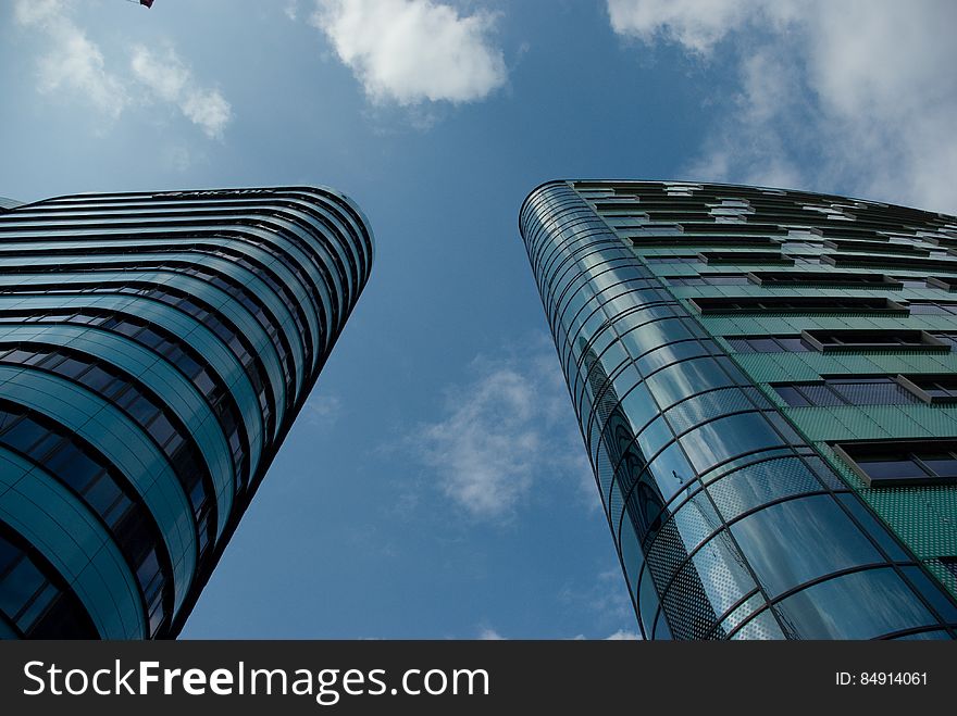 View upwards beside and between two adjacent skyscrapers built mainly of glass and metal, (looking almost identical), background of blue sky and white cloud.