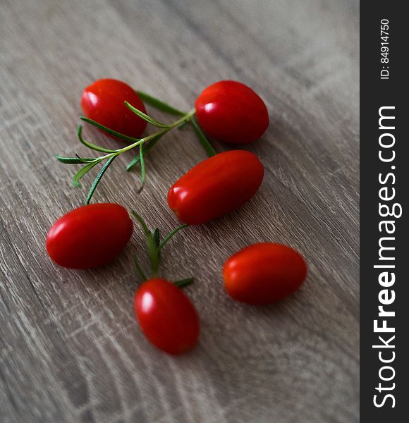 Six small cherry or plum tomatoes together with a sprig of rosemary, lying on a grainy wooden table top. Six small cherry or plum tomatoes together with a sprig of rosemary, lying on a grainy wooden table top.