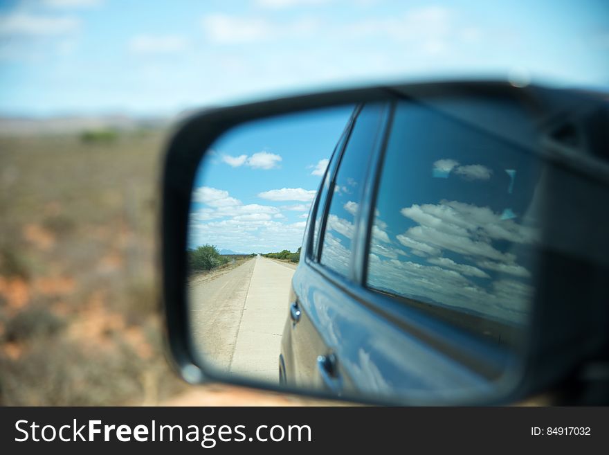 View through car's side mirror showing road, sky, cloud and car windows with blurred view of surrounding countryside. View through car's side mirror showing road, sky, cloud and car windows with blurred view of surrounding countryside.