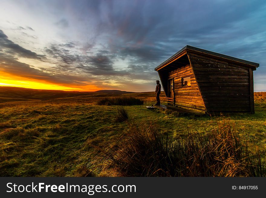 Solitary figure standing beside a log cabin on the moors at sunset with a yellow and orange sky.