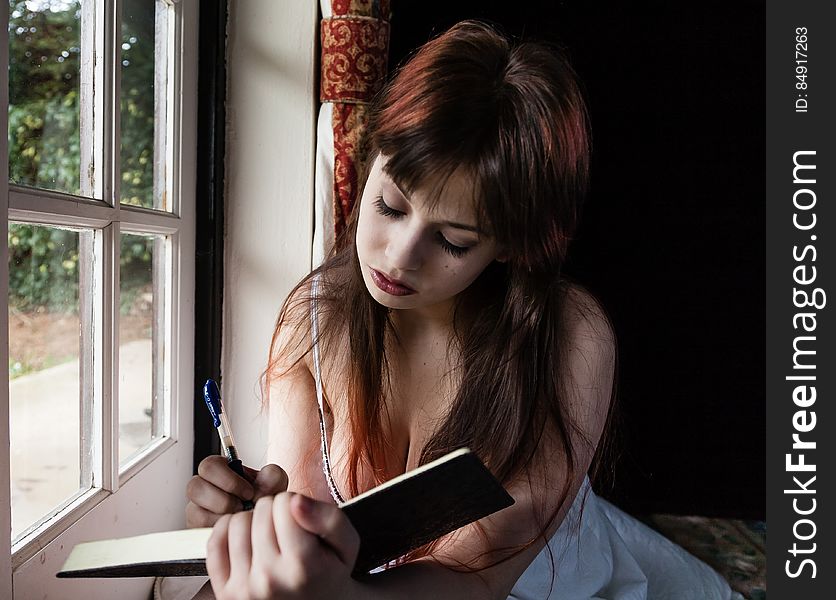 Portrait of pretty young girl with long red hair writing in her diary while seated next to a window. Portrait of pretty young girl with long red hair writing in her diary while seated next to a window.