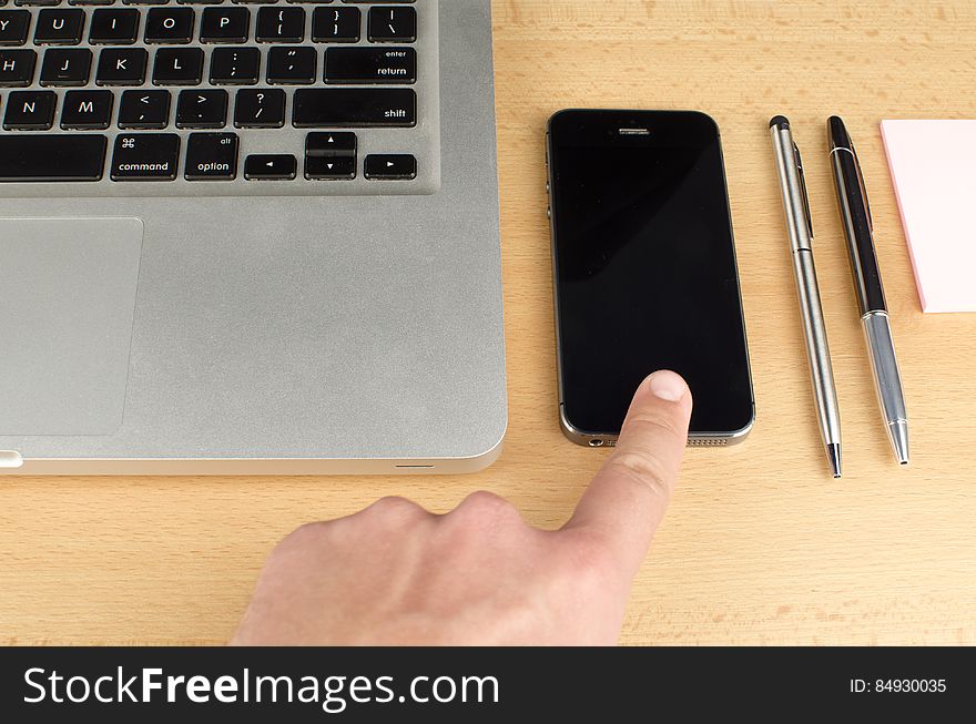 A finger touching a smartphone, laptop keyboard and pens next to it. A finger touching a smartphone, laptop keyboard and pens next to it.
