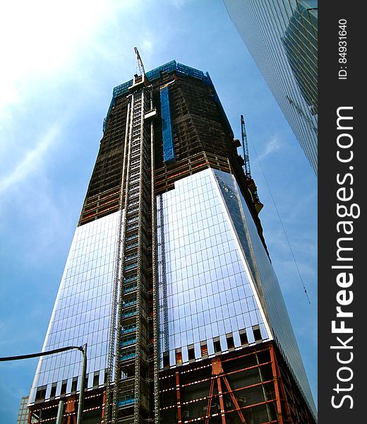 Skyscraper building under construction with ladder (or lift) on the outer wall and heavy lift crane on the far side, blue sky and thin cloud background. Skyscraper building under construction with ladder (or lift) on the outer wall and heavy lift crane on the far side, blue sky and thin cloud background.