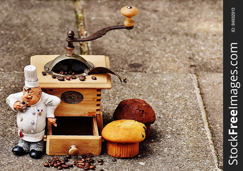 A hand crank coffee bean grinder with muffins and a baker figurine. A hand crank coffee bean grinder with muffins and a baker figurine.