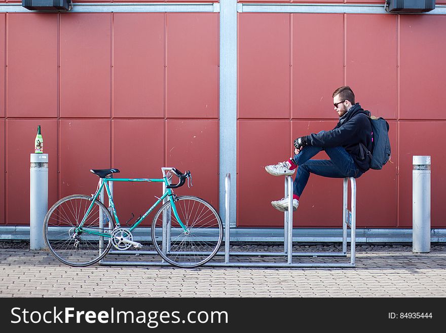 A man sitting on a bike parking rack with a bike parked next to him. A man sitting on a bike parking rack with a bike parked next to him.