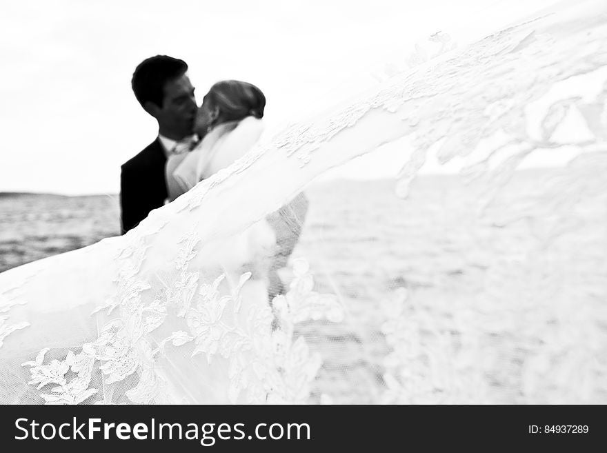 Bride and groom kissing on the sea shore while she is still wearing her dazzling white wedding dress and he a dark formal suit.