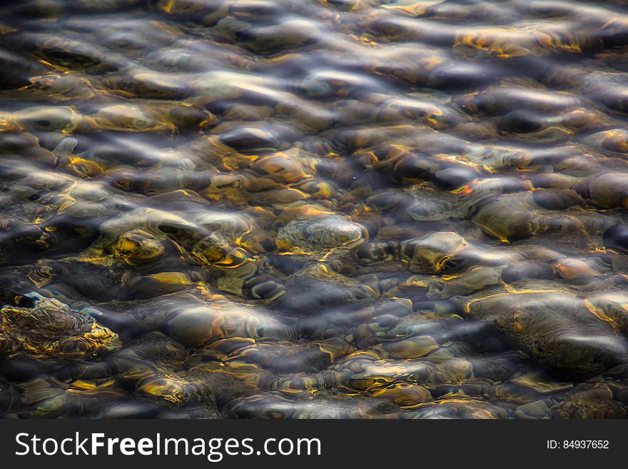 Closeup of shallow ocean waves over rocks and pebbles with golden sunlight reflection at sunset.