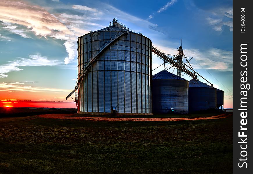 Drying bins for cereal crops on a farm at sunrise with a beautiful red and orange glow and above blue sky and thin clouds. Drying bins for cereal crops on a farm at sunrise with a beautiful red and orange glow and above blue sky and thin clouds.