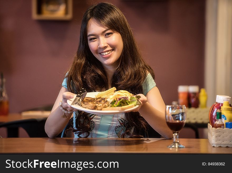 Smiling woman with long brown hair holds out a plate of food (burger or salad rolls) while sitting at a table with a glass of red wine. Smiling woman with long brown hair holds out a plate of food (burger or salad rolls) while sitting at a table with a glass of red wine.