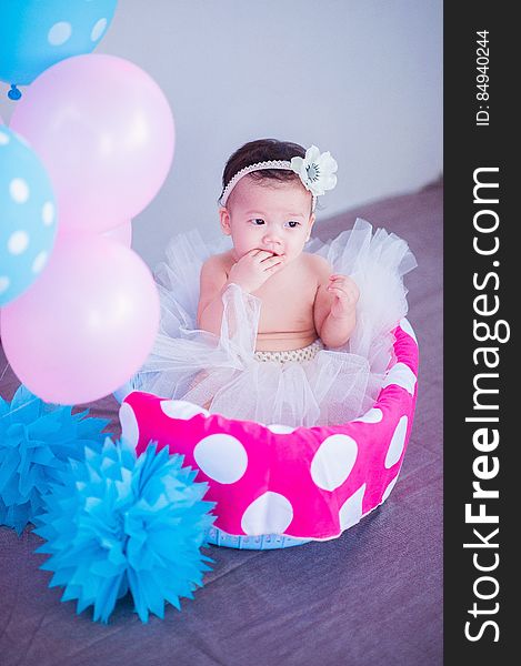 A baby sitting in a basket wearing a tutu with colorful balloons. A baby sitting in a basket wearing a tutu with colorful balloons.