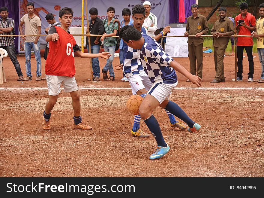 Players on dirt field with spectators during soccer match.