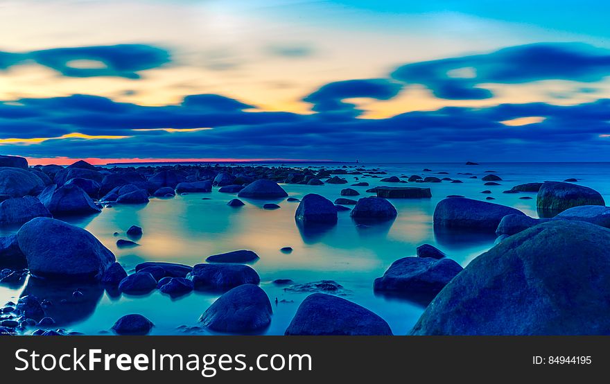 Rocky beach at dusk, sky colors and clouds reflecting on the water surface.
