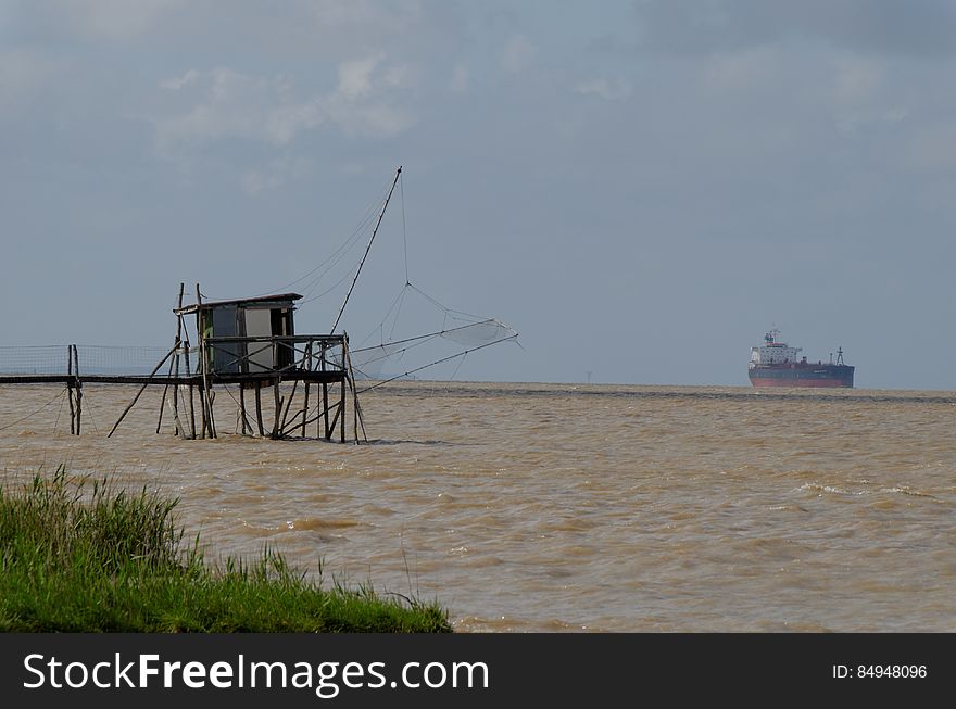 Wooden hut on dock in choppy waters with cargo ship on horizon.