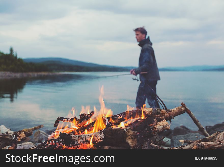 Man Holding Fishing Rod On Lake By Campfire