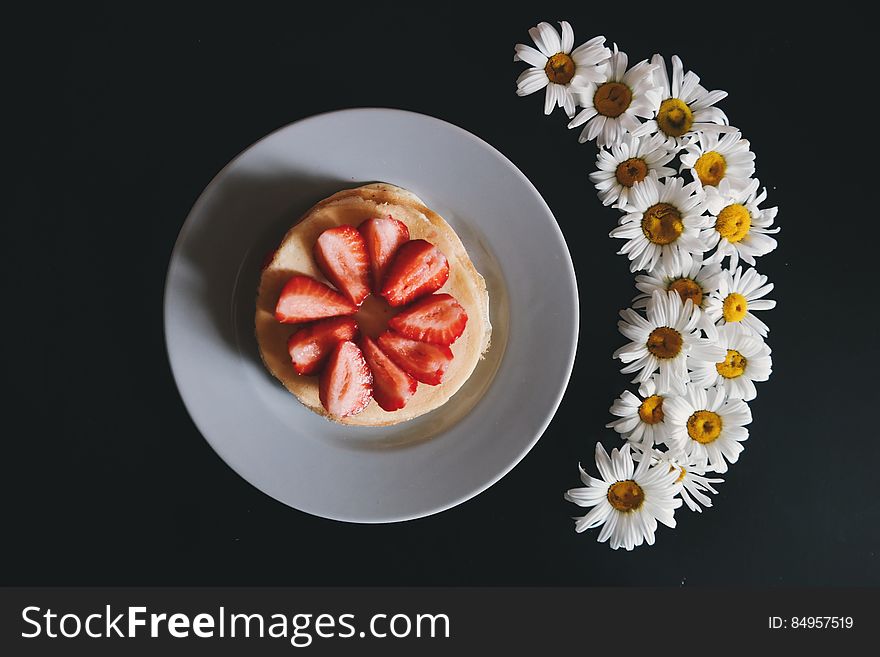 Overhead view of strawberry slices on stack of pancakes next to arranged daisy flowers, black background. Overhead view of strawberry slices on stack of pancakes next to arranged daisy flowers, black background.