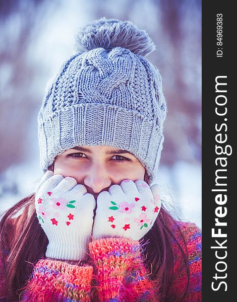 Portrait of pretty young woman in cold Winter weather wearing a gray colored knitted tea cozy hat with pom pom and red and purple jumper with gloved hands covering her mouth, mainly white background. Portrait of pretty young woman in cold Winter weather wearing a gray colored knitted tea cozy hat with pom pom and red and purple jumper with gloved hands covering her mouth, mainly white background.