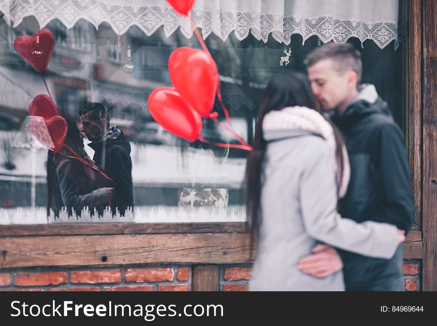 A couple holding heart shaped balloons kissing in front of a window. A couple holding heart shaped balloons kissing in front of a window.