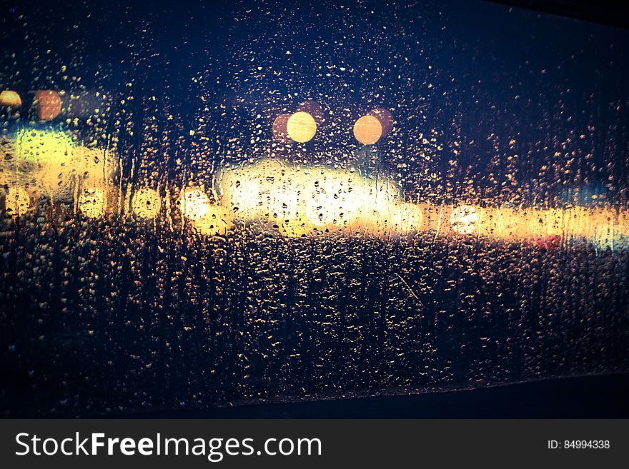 View through a window largely covered in rain drops to a road with traffic and a shopping mall showing up as a gold blur, black night background. View through a window largely covered in rain drops to a road with traffic and a shopping mall showing up as a gold blur, black night background.