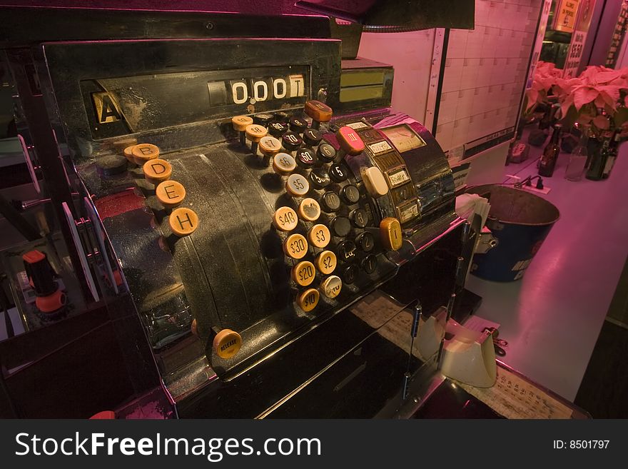 An old soloon cash register, with large push buttons, sits on a counter lit with pink light. An old soloon cash register, with large push buttons, sits on a counter lit with pink light