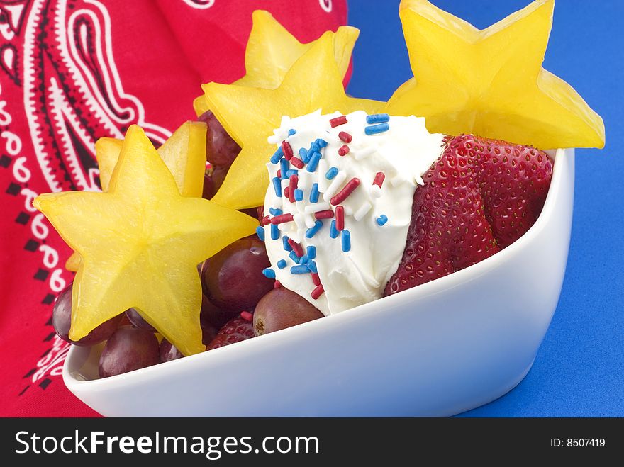 Bowl of colorful starfruit, strawberries and grapes with whipped topping and red, white and blue sprinkles with red bandanna and blue background  copy space. Bowl of colorful starfruit, strawberries and grapes with whipped topping and red, white and blue sprinkles with red bandanna and blue background  copy space