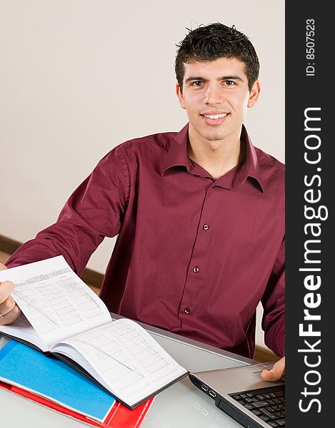 Young man working and smiling at camera with note pad and laptop on desk. Young man working and smiling at camera with note pad and laptop on desk