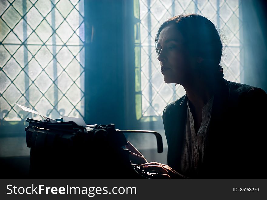 A woman typing on a typewriter by a window. A woman typing on a typewriter by a window.