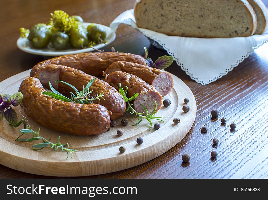 Close up of breakfast sausages with herbs and spices on wooden cutting board with basket of brown break ad bowl of pickles. Close up of breakfast sausages with herbs and spices on wooden cutting board with basket of brown break ad bowl of pickles.
