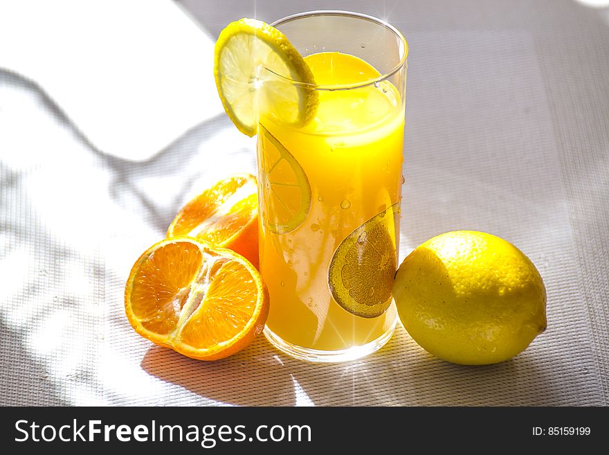 A glass with an yellow drink and lemon slice, lemon and orange next to it. A glass with an yellow drink and lemon slice, lemon and orange next to it.