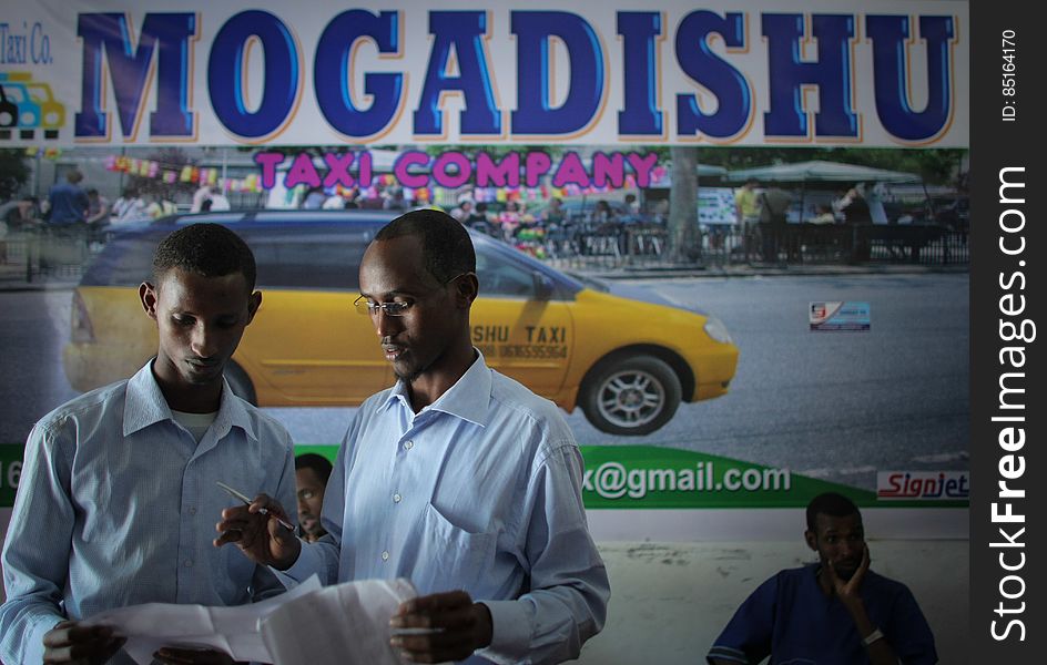 Suleiman Mohamed Daras &#x28;centre&#x29;, manager of the Mogadishu Taxi Company discusses fares and routes with a fellow worker at the company&#x27;s head office in the Somali capital Mogadishu, 01 September 2013. Operating since the end of May of this year in the city&#x27;s dusty and once again bustling streets and thoroughfares, the MTC&#x27;s distinctive yellow and purple cars offer customers taxi fares around Mogadishu at competitive rates and in the first 3 months of operations, the company has increased it&#x27;s fleet of vehicles from an initial 25 to over 100. The company, according to one of it&#x27;s drivers, also enables employment opportunities for Somalia&#x27;s youth following two decades of conflict in the Horn of Africa nation that shattered a generation. Now, thanks to the relative peace that has followed the departure of the Al-Qaeda-affiliated extremist group Al Shabaab from the city; an internationally recognised government for the first time in years and thousands of Diaspora Somalis returning home to invest in and rebuild their country, the MTC is one of many new companies establishing itself in the new Mogadishu and offering services that were hitherto impossible to provide. AU-UN IST PHOTO / STUART PRICE. Suleiman Mohamed Daras &#x28;centre&#x29;, manager of the Mogadishu Taxi Company discusses fares and routes with a fellow worker at the company&#x27;s head office in the Somali capital Mogadishu, 01 September 2013. Operating since the end of May of this year in the city&#x27;s dusty and once again bustling streets and thoroughfares, the MTC&#x27;s distinctive yellow and purple cars offer customers taxi fares around Mogadishu at competitive rates and in the first 3 months of operations, the company has increased it&#x27;s fleet of vehicles from an initial 25 to over 100. The company, according to one of it&#x27;s drivers, also enables employment opportunities for Somalia&#x27;s youth following two decades of conflict in the Horn of Africa nation that shattered a generation. Now, thanks to the relative peace that has followed the departure of the Al-Qaeda-affiliated extremist group Al Shabaab from the city; an internationally recognised government for the first time in years and thousands of Diaspora Somalis returning home to invest in and rebuild their country, the MTC is one of many new companies establishing itself in the new Mogadishu and offering services that were hitherto impossible to provide. AU-UN IST PHOTO / STUART PRICE.