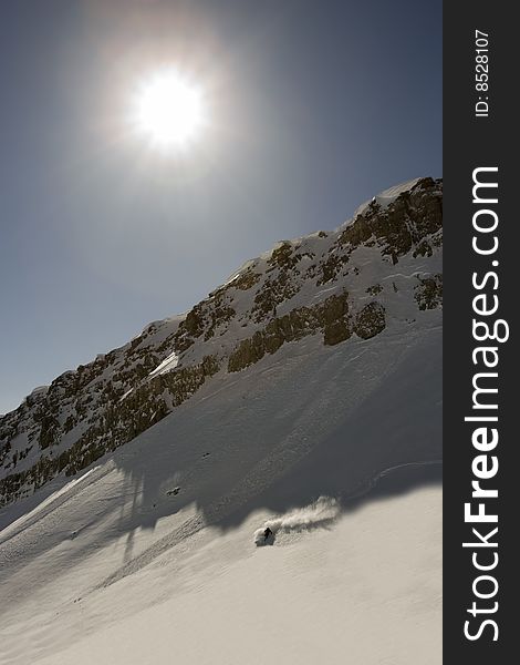 A lone Skier turns in powder under rock ridge and bright sun. A lone Skier turns in powder under rock ridge and bright sun