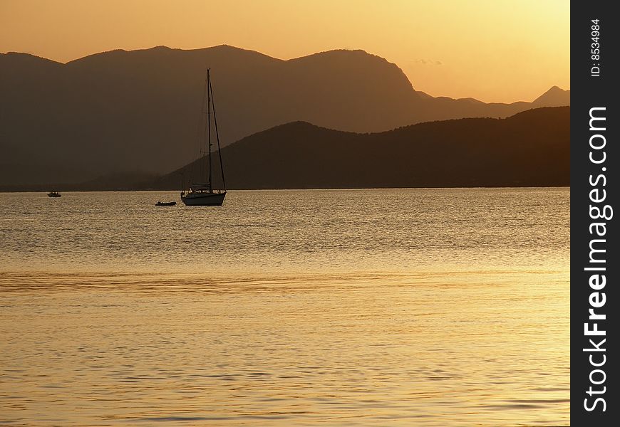 The sun sets over a sailboat on the sea with mountains as a background. The sun sets over a sailboat on the sea with mountains as a background