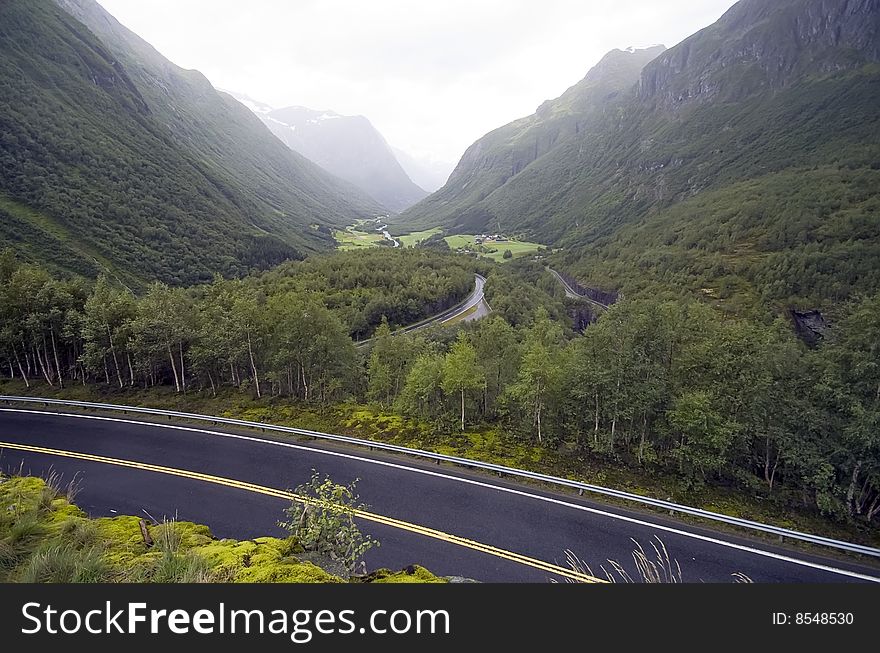 A valley between forest covered mountains. Norwegian landscape on a rainy and misty day. Wet road in the foreground. A valley between forest covered mountains. Norwegian landscape on a rainy and misty day. Wet road in the foreground.