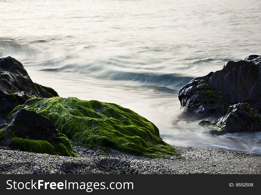 Long exposure on shore, blurred waves over green rocks.