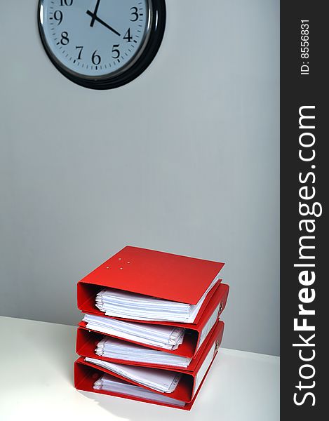 Stack of red folders on desk and a clock in background, deadline concept