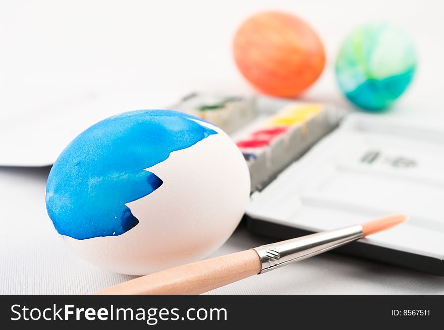 A half painted blue easter egg with brush and watercolor palette and color easter eggs in background. A half painted blue easter egg with brush and watercolor palette and color easter eggs in background.