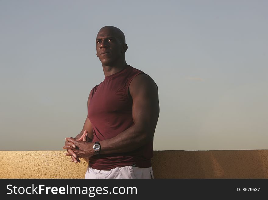 A male athlete with dark skin wearing a puple shirt relaxes out doors against a mustard wall and blue sky. A male athlete with dark skin wearing a puple shirt relaxes out doors against a mustard wall and blue sky