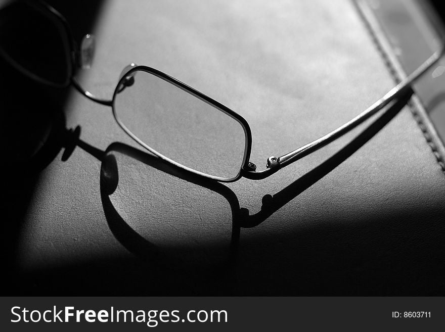 Pair of man's glasses on a leather business pad silhouetted against the sun. Sharp shadow on left hand side. Pair of man's glasses on a leather business pad silhouetted against the sun. Sharp shadow on left hand side